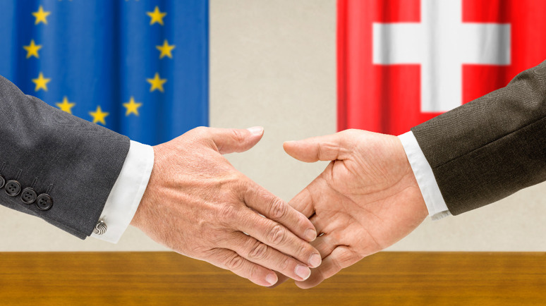 Due mani si stringono. Sullo sfondo, una bandiera dell'UE e una della Svizzera